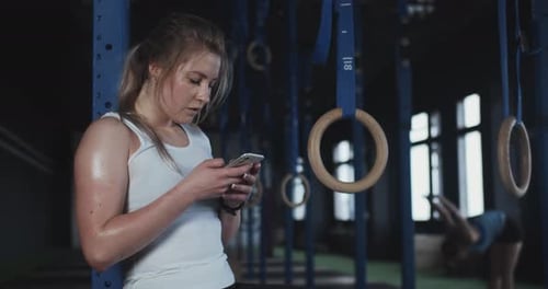 Woman Using Phone in Gym Near Rings
