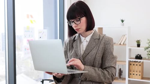 Beautiful Woman in Suit Standing with Portable Computer Near Office Table
