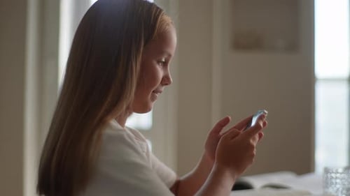 Side View of Pretty Child Girl 10s Sitting at Home Table Alone with Cellphone Having Fun on Internet