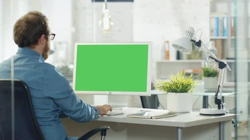 Young Man with a Beard Actively Works on His Desktop Computer. He Sits In His Modern Studio/Office.
