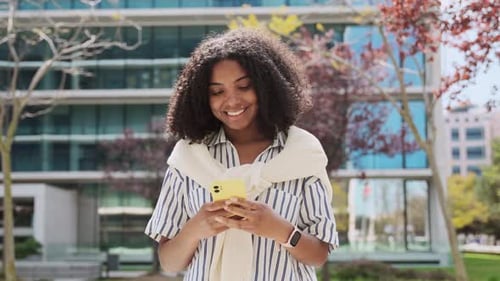 Young Happy African American Woman Walking in City Park Using Mobile Phone