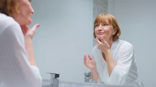 Smiling Woman Applying Lotion in Bathroom Mirror