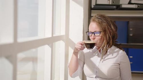 Successful business woman enjoys drinking coffee and looking out window on sunny morning in office
