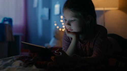 Jolie petite fille dans sa chambre la nuit, allongée sur un lit avec une tablette informatique. Sa lampe de nuit s'est allumée