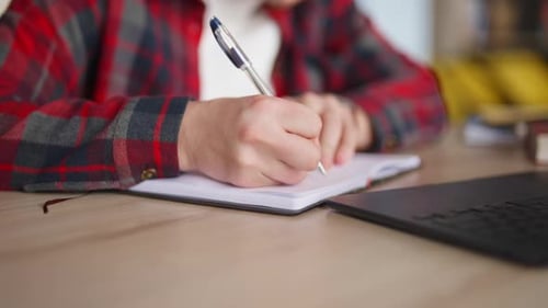 Adult Male Writing in Notebook Beside Laptop Indoors