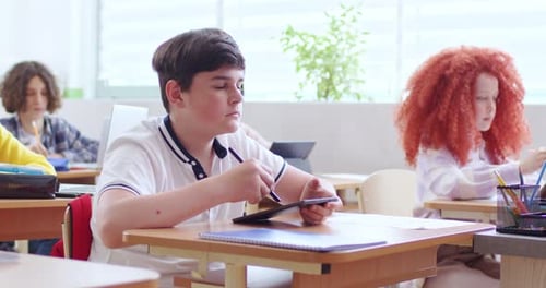 Teen Caucasian Schoolboy Sitting at Desk in Classroom Among Kids at Lesson and Using Tablet Device