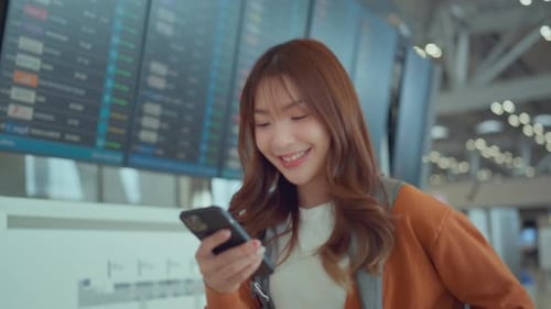 Happy asian woman traveller checking flight schedule departures board in airport terminal