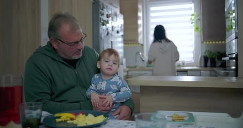Father holding baby at dining table, baby gazing upwards while mother works in kitchen, warm family