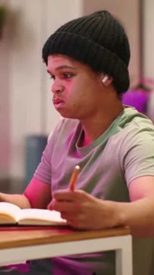 Vertical video: Shifting to focused man at table after glance, writing in notebook with pen, beanie
