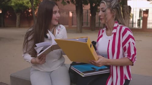Zwei junge Frauen, wahrscheinlich Studenten oder Gymnasiasten, sitzen draußen auf einer Bank und teilen sich Notizen und