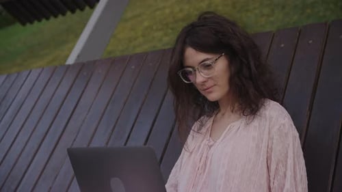 Handheld Close Up of a Curly Caucasian Woman Working on Her Laptop Computer on a Park Bench