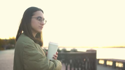 Young Woman with a Glass of Coffee in Her Hand A Girl in Glasses Near the Fence on the Embankment