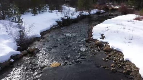 Small stream with fresh snow on the banks and trees in the background. Moving drone shot following