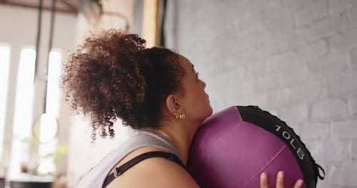Medicine ball descending toward woman catching, squatting, propelling ball against wall for workout