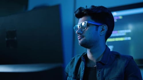 Konzentrierter junger männlicher Softwareentwickler mit Brille beim Programmieren spät in der Nacht in einem dunklen Büro.