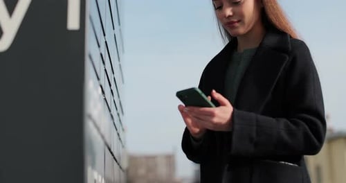 Woman Receiving Parcel From Post Terminal Machine Using Smartphone Outdoors Parcel Delivery Machine