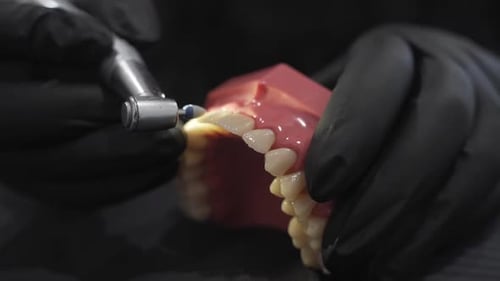 Close-up of a dental drill polishing a set of prosthetic teeth held by gloved hands