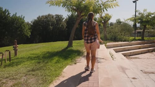 Woman walks along a Brick Pathway beside Green Shrubs and a Tree, with Steps leading to a Sunlit Pla