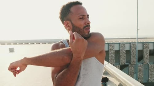 Young athlete doing warm-up, stretching his arms before training while standing on the embankment