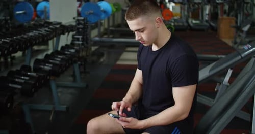Portrait of Guy in Gym While Resting Between a Workout Uses Phone Body Strength Fit Health Male