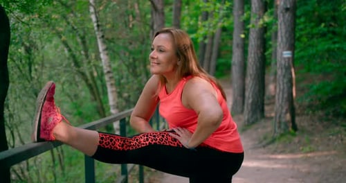 A Caucasia lady is in sportswear doing stretching exercises in a park surrounded by woods.