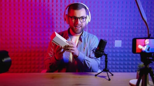 Young Smiling Blogger Unpacking Box with Present Sitting at Home Studio