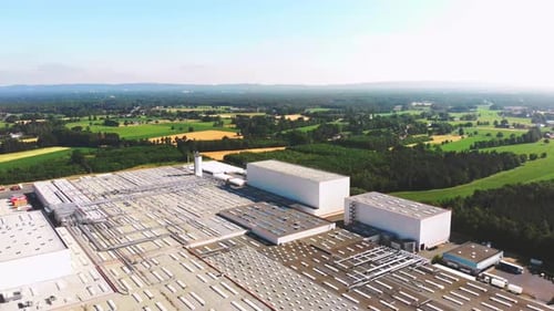 Aerial wide shot of a large industrial warehouse complex surrounded by green farmland and forests