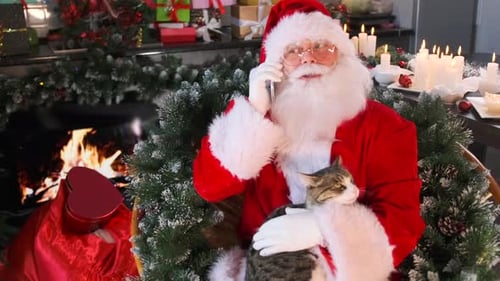 Le Père Noël est assis en train de caresser un chat dans sa résidence près de la cheminée et discute joyeusement sur son téléphone
