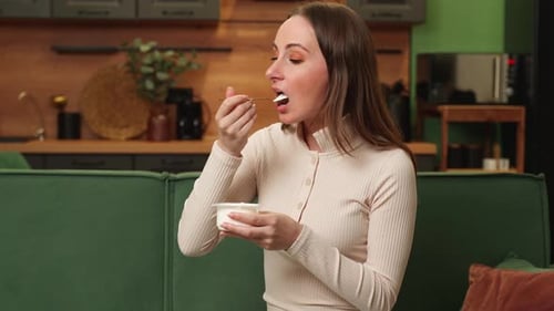 Happy Brunette Woman Eats Delicious Yogurt at Home Sitting on a Green Sofa