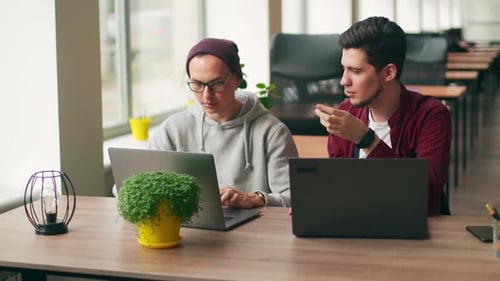Two Hipster Coworkers Sharing Ideas About Project Sitting with Laptops in Spacious Open Space Office