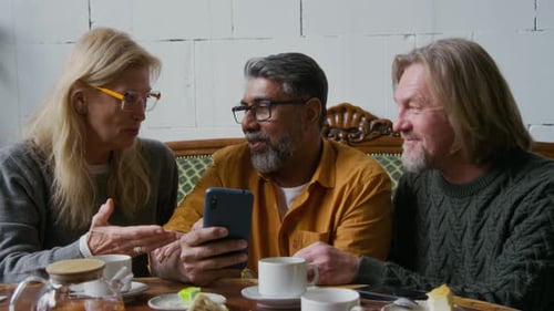 Cheerful Mature Friends Laughing at Photos on Smartphone in Cafe