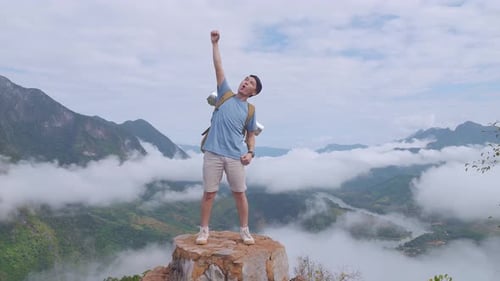 Männlicher Wanderer, der auf dem Felsen steht und seine Hand hebt und feiert, den Gipfel des nebligen Berges zu erklimmen