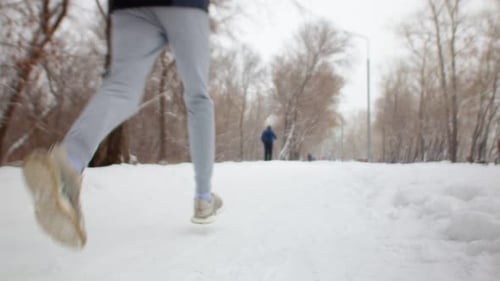 Slow Motion View From Bottom Behind a Man in Sports Clothes Running in a Snowy Winter Park Snow is