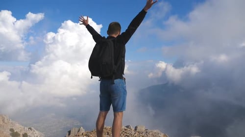 Man Tourist with Backpack Standing on the Edge of the Hill and Victoriously Outstretching Arms Young