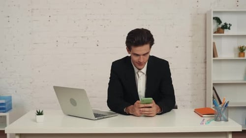 Medium Video of a Young Man Sitting in the Office at the Table with a Smartphone in His Hands