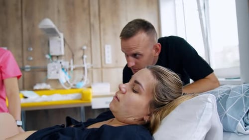 Mujer caucásica cansada yace en la cama lista para dar a luz. El marido está de pie junto a su cabeza hablando con ella.