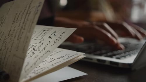 An asian woman types on her laptop keyboard out of focus while a notebook with notes flaps in the wi