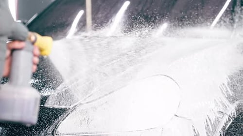 close-up A male car wash employee applies car wash detergent to a black luxury car using spray gun