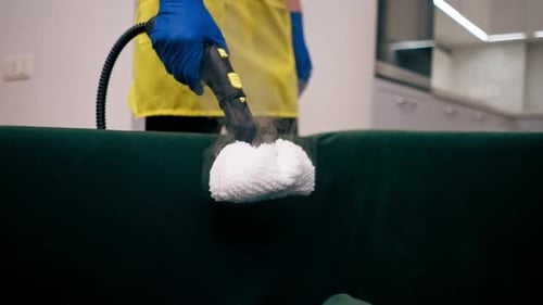 Close Up in an Apartment Cleaner in a Yellow Apron Does a Steam Cleaning of a Green Sofa with