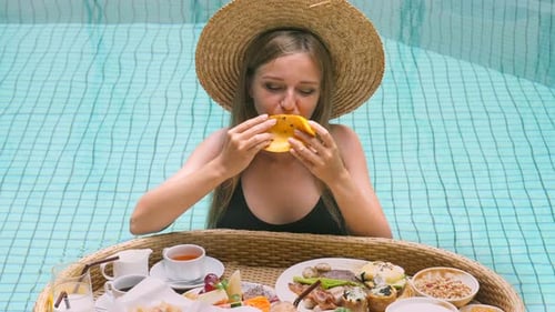 Woman in Swimming Pool with Floating Breakfast Eating Delicious Mango