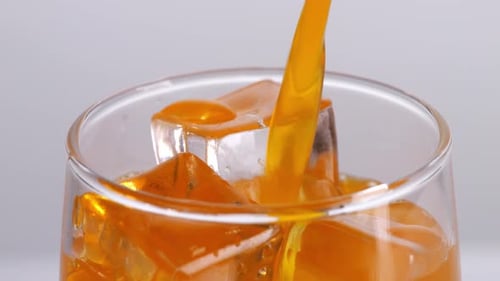 Close Up Of Orange Juice Being Poured Into A Glass With Ice Cubes On The Light White Background