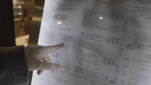 Hand pointing at a menu outside a Paris café, showing French food and drinks options