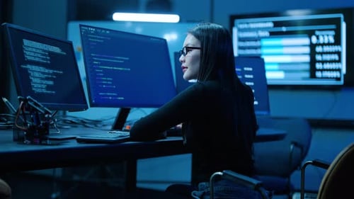 Focused female programmer coding on multiple monitors in a dark high-tech cybersecurity center.