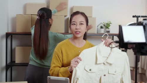 Young asian woman live online clothing store while showing clothing with tablet on social at home.