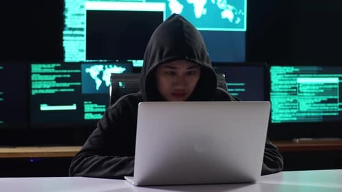 Eine Hackerin in einer Kapuze arbeitet an einem Laptop mit Daten auf Bildschirmen mehrerer Monitore