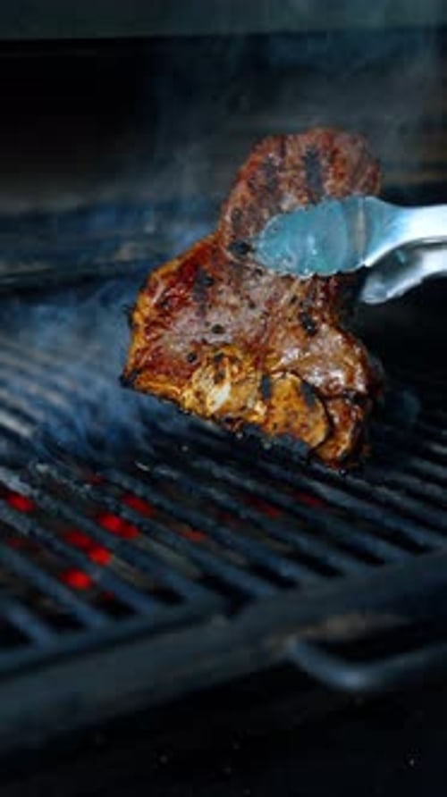 Ein Stück Rindfleisch mit Knochen wird auf dem Grill gegart. Fleisch wird mit der Zange gedreht. Nahaufnahme.