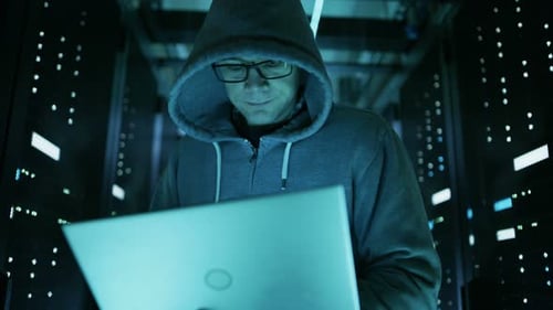 Low Angle Shot of Computer Hacker in Hoodie, Holding Laptop. He Breaked in a Corporate Data Center.