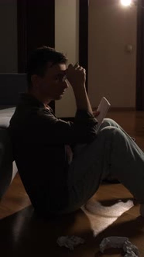 Vertical Video Young Man Sitting on the Floor Holding a Pen and Paper Contemplating Ideas