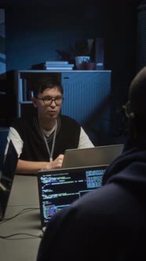 Probador de software conversando con un desarrollador web que trabaja en computadoras portátiles en una oficina de TI oscura