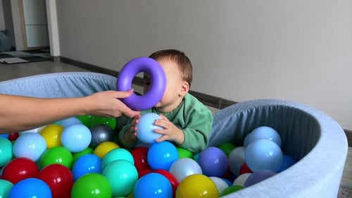 Happy Caucasian boy lay on the wall of the soft dry basin. Mom’s hand gives a toy ring to her son.